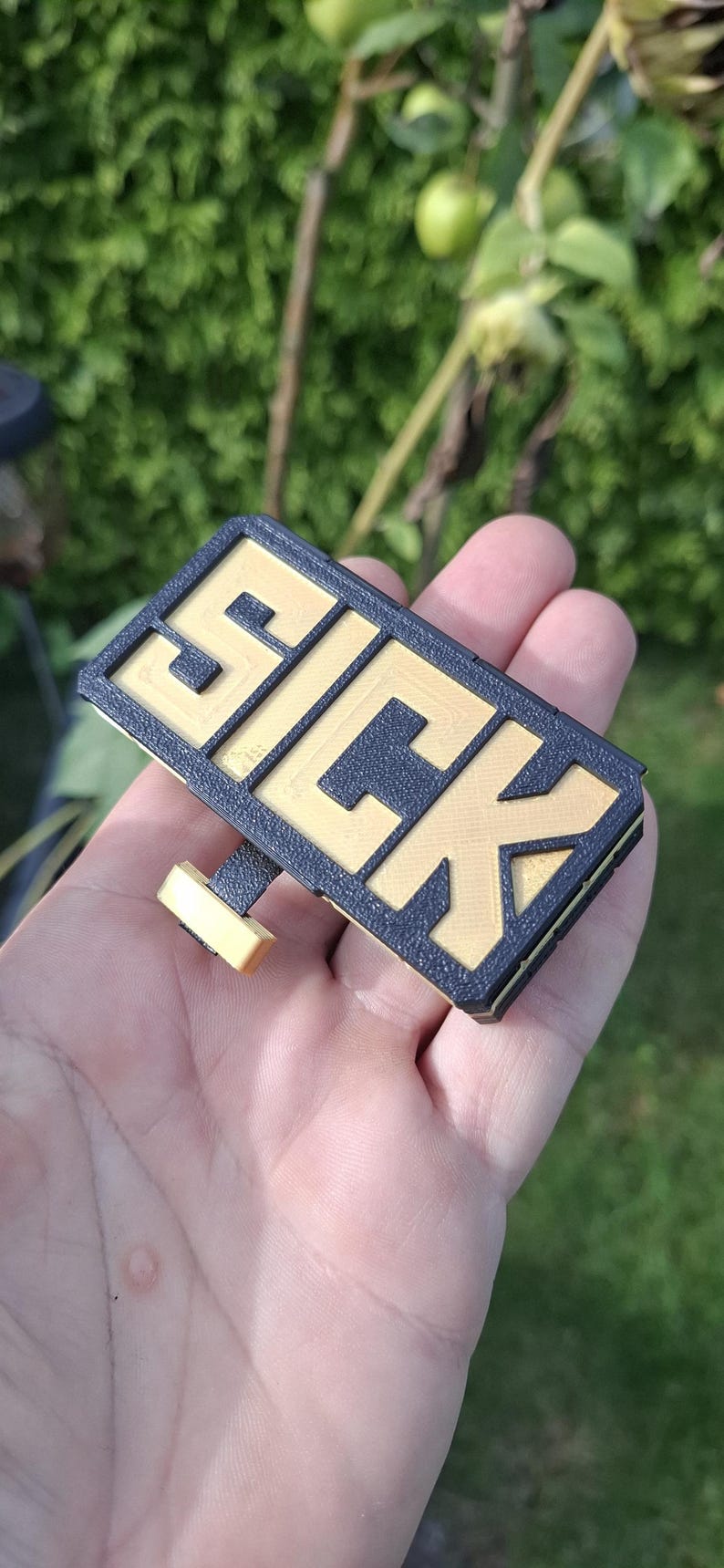 May include: A rectangular 3D printed sign featuring the word SICK in raised gold lettering against a textured black background. The object includes a small gold and black attachment extending from the bottom edge, showcasing a layered construction.