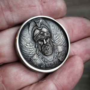 Puede incluir: Una moneda de plata con una escultura en relieve detallada de un guerrero alado que lleva un casco y una armadura. El guerrero tiene barba y una expresi&oacute;n seria.