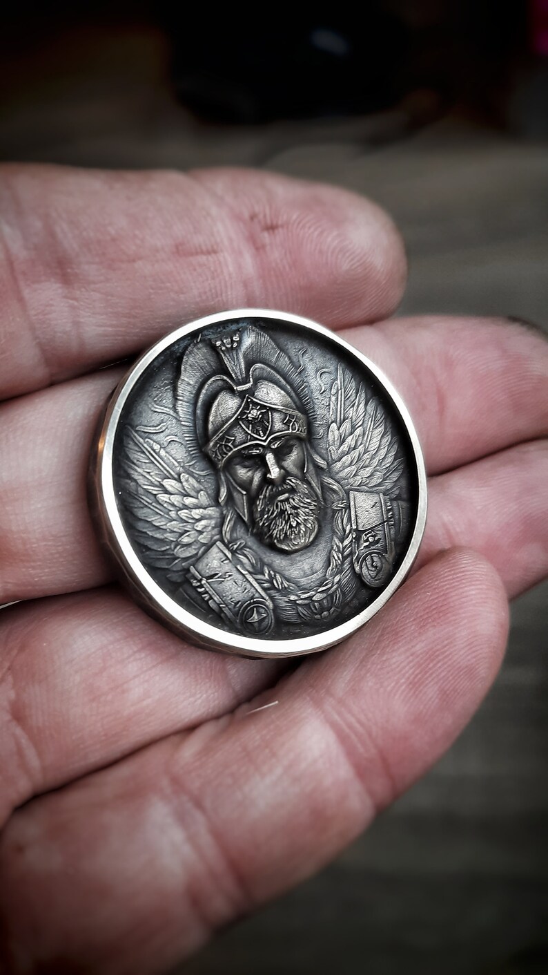 Puede incluir: Una moneda de plata con una escultura en relieve detallada de un guerrero alado que lleva un casco y una armadura. El guerrero tiene barba y una expresi&oacute;n seria.
