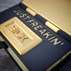 May include: A black and gold rectangular object with a hinged design. The surface features the embossed text JUST FREAKIN' in gold lettering, with a raised gold rectangular plaque displaying the word SICK in bold, three-dimensional letters.