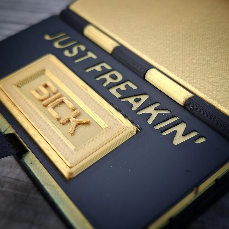 May include: A black and gold rectangular object with a hinged design. The surface features the embossed text JUST FREAKIN' in gold lettering, with a raised gold rectangular plaque displaying the word SICK in bold, three-dimensional letters.