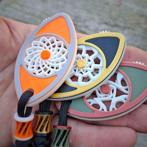 May include: Three eye-shaped fidget spinners with intricate white geometric centers. The outer frames are orange, black, and olive green, each featuring a matching colored center button and a black cord with a decorative matching bead at the base.