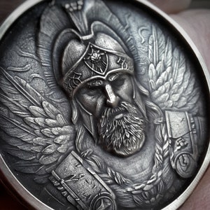 Puede incluir: Una moneda de plata con una escultura en relieve detallada de un guerrero alado que lleva un casco con una cresta. El guerrero tiene barba y una expresi&oacute;n seria.