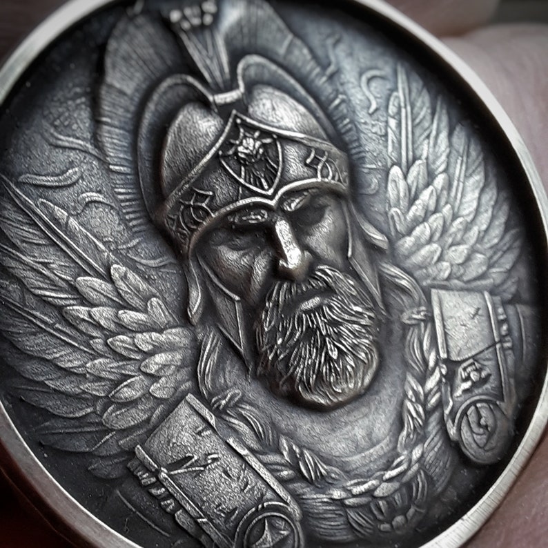 Puede incluir: Una moneda de plata con una escultura en relieve detallada de un guerrero alado que lleva un casco con una cresta. El guerrero tiene barba y una expresi&oacute;n seria.