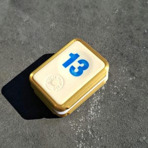 Peut inclure: Un carreau blanc avec une bordure dorée et le numéro "13" imprimé en bleu. Le carreau a un petit logo circulaire avec le mot "SUNRISE" imprimé dessus.