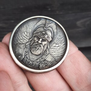 Puede incluir: Una moneda de plata con una escultura en relieve detallada de un guerrero que lleva un casco y una armadura. El guerrero tiene barba y alas en la espalda.