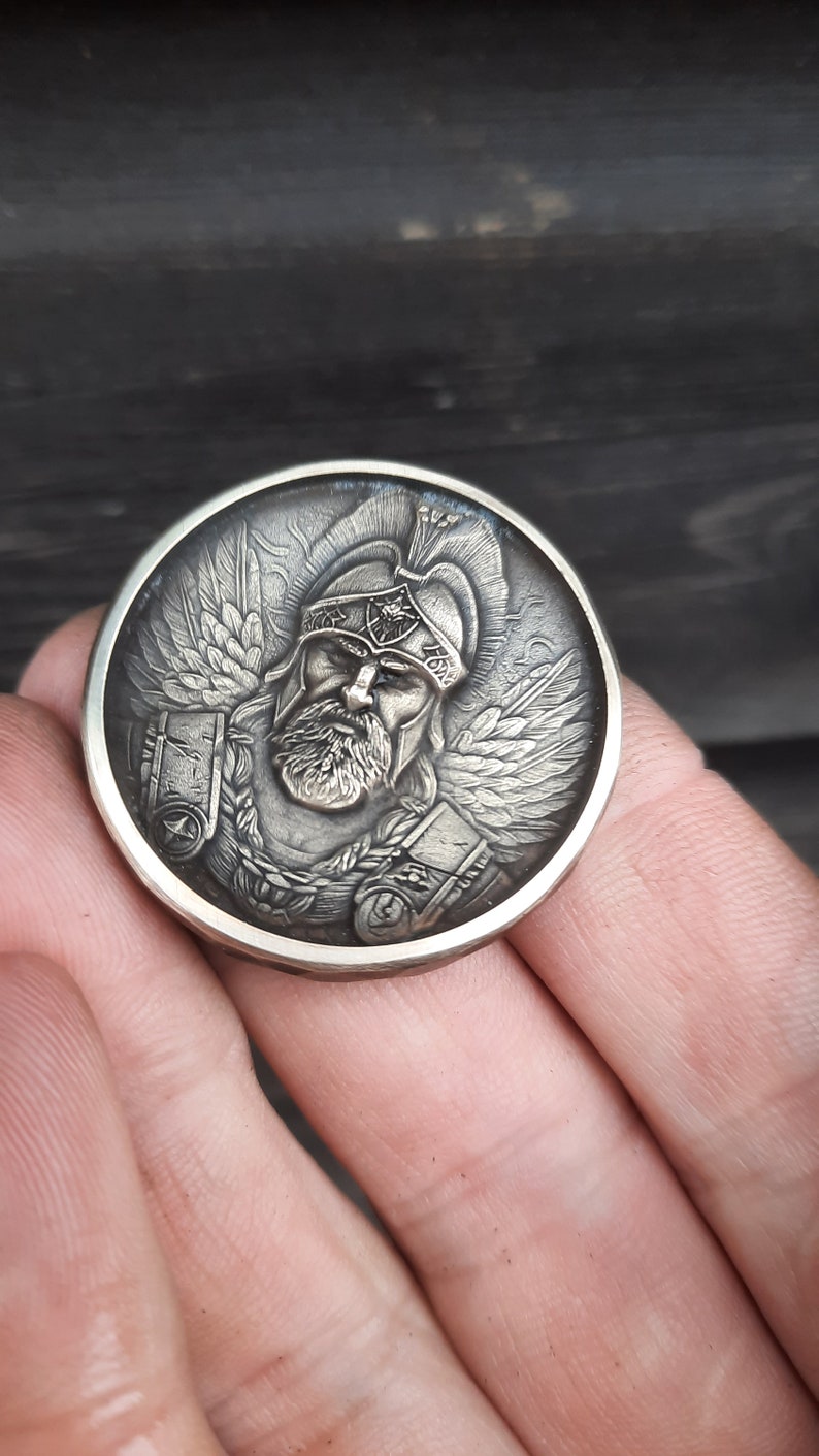 Puede incluir: Una moneda de plata con una escultura en relieve detallada de un guerrero que lleva un casco y una armadura. El guerrero tiene barba y alas en la espalda.