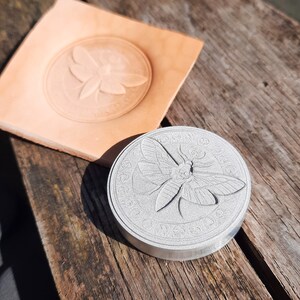 May include: A silver metal stamp with a detailed design of a moth with intricate wings. The stamp is circular and has a raised edge.