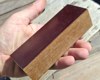 May include: A rectangular block with a dark maroon top surface featuring a fine cross-hatched texture and a light brown, porous, fibrous side panel. The object has sharp, clean edges and a solid, dense appearance, showing a clear contrast between the smooth, dark top and the rough, tan-colored side.