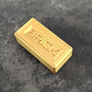 May include: A rectangular, gold-coloured object with a textured surface and the word BRICK embossed in capital letters on the top face. The item features a layered, block-like construction with visible horizontal lines along the sides and edges.