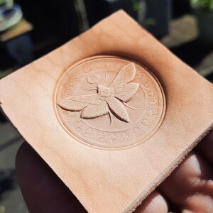 May include: A close-up of a leather stamp with a detailed design of a moth with wings spread. The stamp is circular and has a raised border.