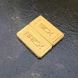 May include: Two rectangular, gold-coloured plastic tiles featuring raised lettering. The top tile displays the word SICK, while the bottom tile displays the word BRICK. Both tiles have a textured, lined surface within a raised border frame.