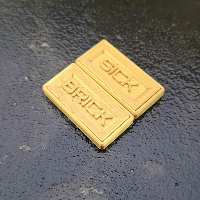 May include: Two rectangular, gold-coloured plastic tiles featuring raised lettering. The top tile displays the word SICK, while the bottom tile displays the word BRICK. Both tiles have a textured, lined surface within a raised border frame.