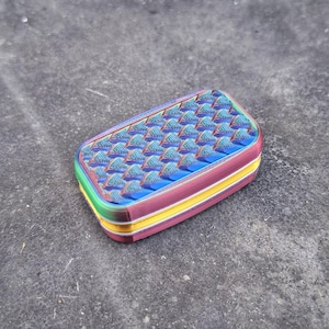 May include: A rectangular, multi-colored object with a textured surface. The top features a pattern of small, blue and purple pyramid shapes. The sides are layered with green, yellow, and maroon bands. The object has rounded corners.