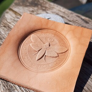 May include: A light brown leather coaster with a circular design featuring a detailed embossed image of a moth with intricate wings and a circular border.