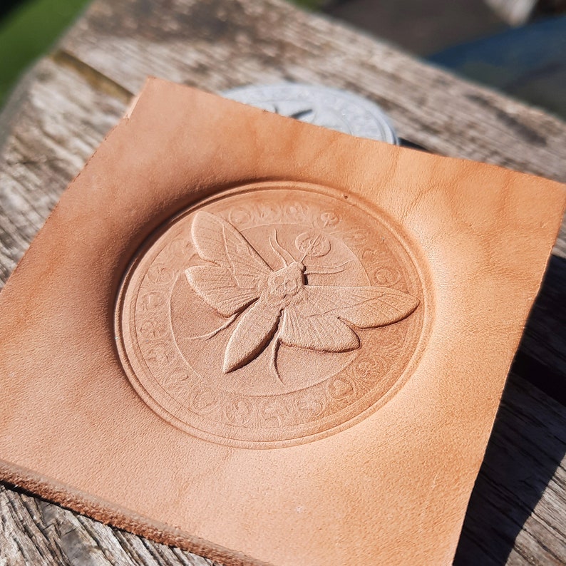 May include: A light brown leather coaster with a circular design featuring a detailed embossed image of a moth with intricate wings and a circular border.