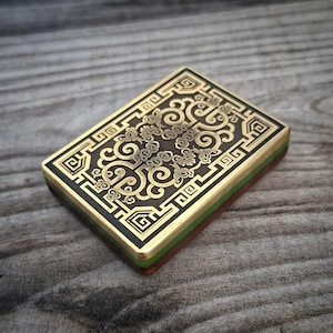 Pode incluir: Um objeto retangular com uma placa superior de metal dourado apresentando intrincados gravados pretos de volutas e bordas geométricas. A base consiste numa estrutura de camadas com uma secção central verde e uma camada inferior castanha.