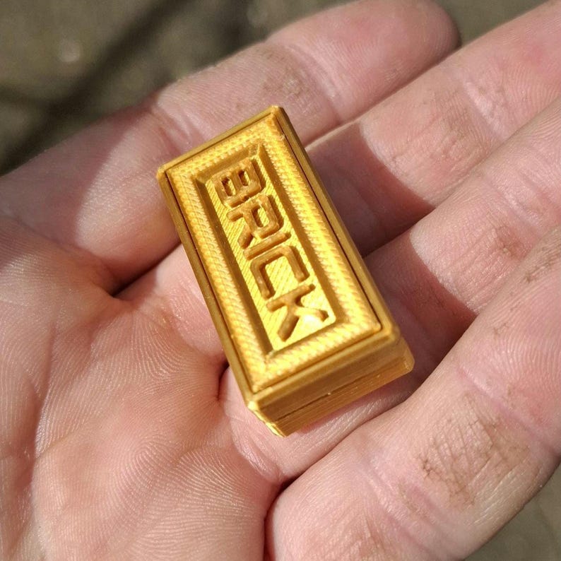 May include: A small, rectangular, gold-coloured 3D printed object with a textured surface. The top face features a raised border and the word BRICK embossed in capital letters in the centre. The object has a layered construction visible on its sides.