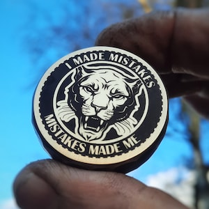 Puede incluir: Un token redondo de metal negro y dorado con un gráfico de cabeza de puma y el texto "I Made Mistakes Mistakes Made Me".