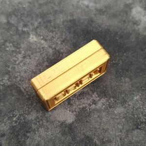 May include: A rectangular gold-coloured object with a smooth top surface and a textured, layered side profile. The item features a distinct horizontal structure with visible linear ridges along the edges, presenting a solid, block-like form with a metallic finish.