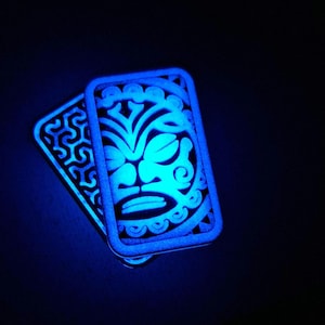 Op de afbeelding: Twee rechthoekige, lichtgevende blauwe objecten met ingewikkelde uitgesneden patronen. Het bovenste object heeft een gestileerd, symmetrisch gezicht met gebogen lijnen, terwijl het onderste object een herhalend, geometrisch vlechtpatroon toont.