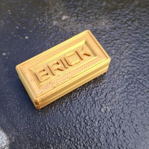 May include: A rectangular, gold-coloured plastic brick with the word BRICK embossed in capital letters on the top surface. The object features a layered, textured finish consistent with 3D printing, showing horizontal lines along the sides.