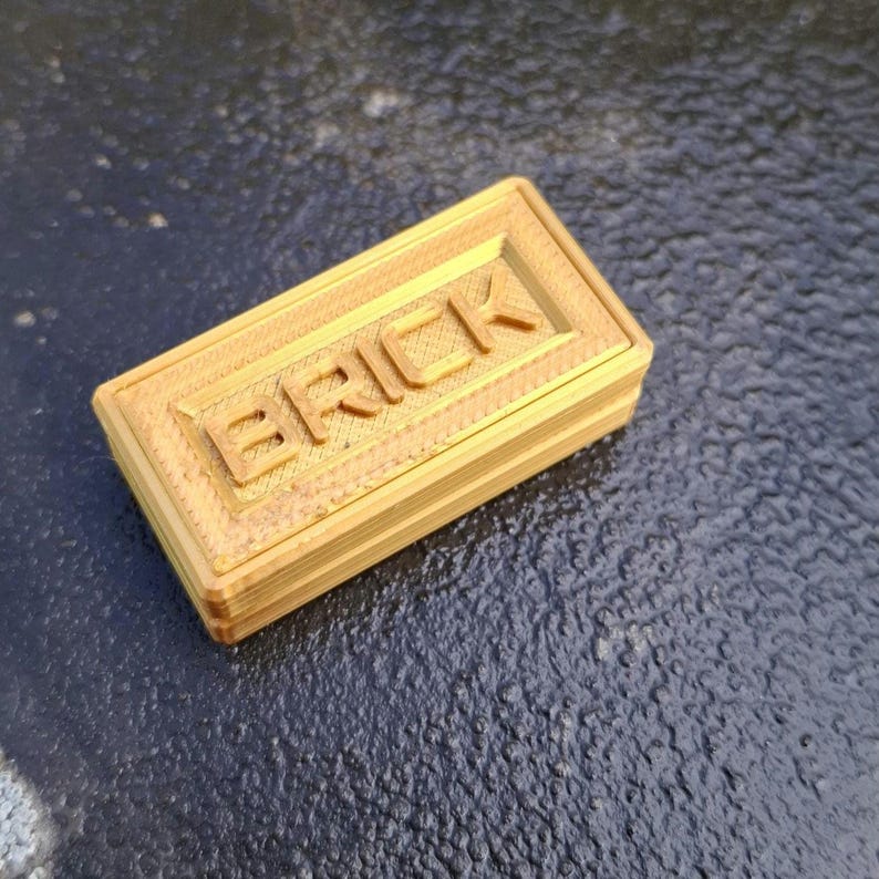 May include: A rectangular, gold-coloured plastic brick with the word BRICK embossed in capital letters on the top surface. The object features a layered, textured finish consistent with 3D printing, showing horizontal lines along the sides.