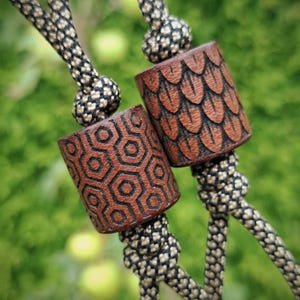 May include: Two cylindrical brown wooden beads threaded onto black and tan paracord. One bead features a carved hexagonal geometric pattern, while the other displays a textured scale-like design. Both beads are secured with knots.