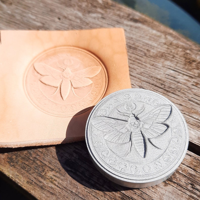 May include: A silver metal stamp with a detailed design of a moth with wings spread, surrounded by a circle of text. The stamp is used to create an impression on leather.