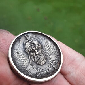 Puede incluir: Una moneda redonda plateada con una escultura en relieve detallada de un guerrero alado con casco. El guerrero tiene barba y una expresi&oacute;n seria.