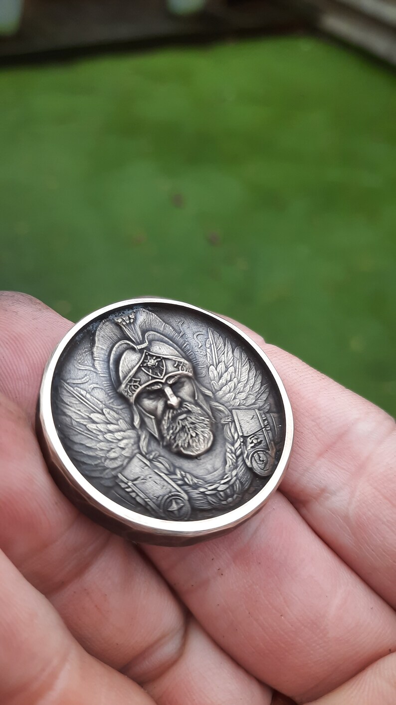 Puede incluir: Una moneda redonda plateada con una escultura en relieve detallada de un guerrero alado con casco. El guerrero tiene barba y una expresi&oacute;n seria.