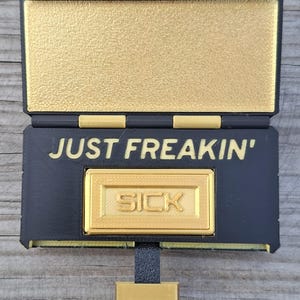 May include: A black and gold rectangular hinged box with a textured finish. The interior lid is gold, while the base features the black text "JUST FREAKIN'" above a raised gold rectangular plaque with the word "SICK" embossed in the centre.