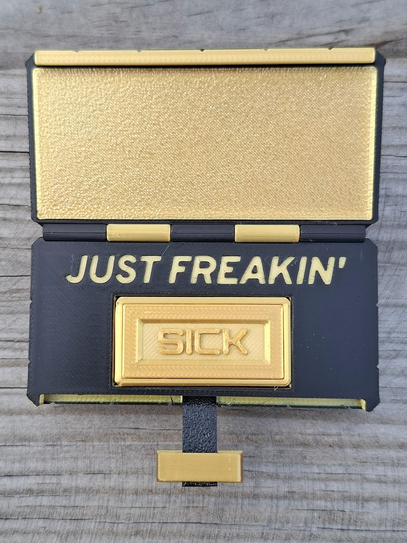 May include: A black and gold rectangular hinged box with a textured finish. The interior lid is gold, while the base features the black text "JUST FREAKIN'" above a raised gold rectangular plaque with the word "SICK" embossed in the centre.
