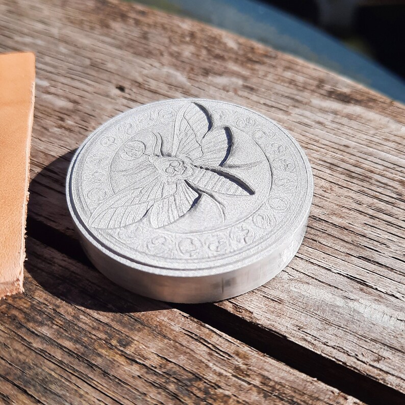 May include: A silver-colored metal stamp with a detailed design of a moth with wings spread. The moth is surrounded by a circular pattern of decorative elements.
