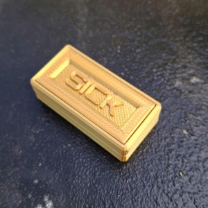 May include: A rectangular gold-coloured 3D printed box with a textured surface. The lid features the word SICK embossed in large capital letters, surrounded by a cross-hatched grid pattern within a raised border frame on the top surface.