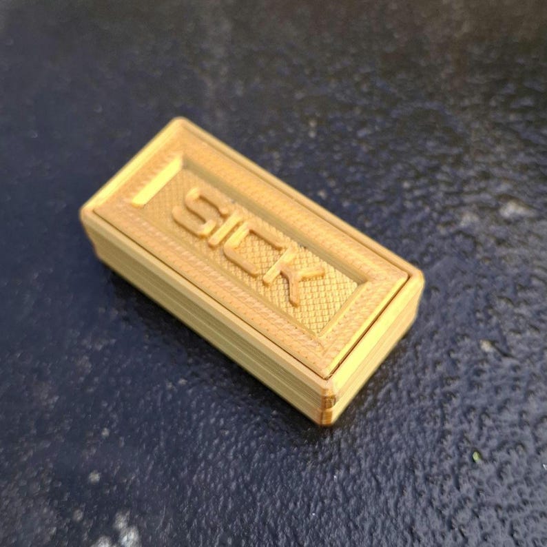 May include: A rectangular gold-coloured 3D printed box with a textured surface. The lid features the word SICK embossed in large capital letters, surrounded by a cross-hatched grid pattern within a raised border frame on the top surface.
