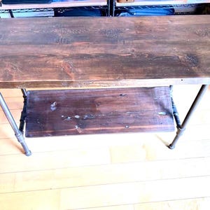 May include: A rustic wooden desk with a dark brown tabletop and a lower shelf. The desk has metal legs and a simple, industrial design. The wood grain is visible, adding to its natural aesthetic. The desk is suitable for home or office use.