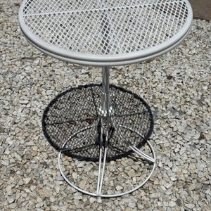 May include: A white, round metal patio table with a mesh top and a circular base. The table has a single central support and is set on a bed of small stones. The table's diameter is approximately 24 inches.