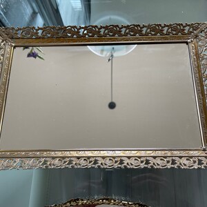 May include: A gold-colored metal tray with a decorative, ornate border. The tray has a mirrored surface.