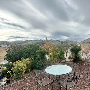 May include: A glass-top patio table with four black metal chairs sits on a bed of brown wood chips. The table and chairs are arranged in a square formation. The background shows a view of rolling hills and a cloudy sky.