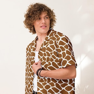 May include: A short-sleeved button-down shirt with a giraffe pattern in shades of brown and white. The shirt has a classic collar and is worn open over a white undershirt. The model is wearing a black bracelet.