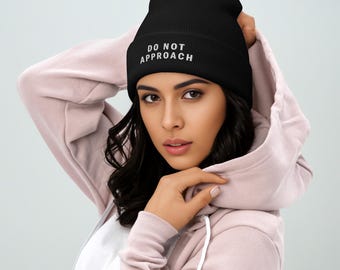 May include: Black beanie with the embroidered text "DO NOT APPROACH" in white. The beanie is worn by a person wearing a pink hooded sweatshirt.