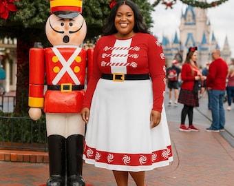 Puede incluir: Una mujer con un vestido rojo y blanco con cinturón negro y hebilla dorada, zapatos rojos y un top rojo a juego. Un gran soldado de madera está cerca. El fondo incluye un árbol de Navidad y un castillo.