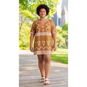 May include: A brown, short-sleeved dress with a gingerbread theme. The dress features candy cane and gingerbread man designs in red, white, and green. The dress is worn with white sandals. The background includes a wooden walkway and a park.