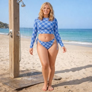 May include: A blue and white plaid long-sleeve crop top and matching bikini bottoms. The swimwear is worn on a sandy beach with the ocean in the background. A wooden shower fixture is on the left.