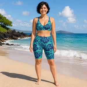 May include: A teal two-piece athletic set featuring a floral pattern in dark blue. The set includes a sports bra and biker shorts. The woman is standing on a sandy beach with the ocean in the background.
