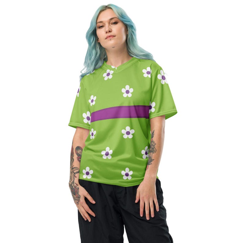 Puede incluir: Una camiseta verde lima con cuello en V y mangas cortas. La camiseta presenta un estampado de flores blancas con centros morados y una franja horizontal morada en el pecho. La persona lleva pantalones negros.