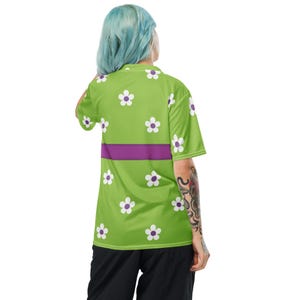 Puede incluir: Una camiseta verde lima de manga corta con estampados de flores de margarita blancas y una franja horizontal p&uacute;rpura. La camiseta est&aacute; hecha de un material ligero.