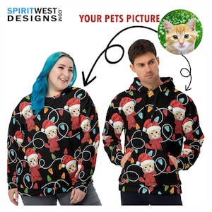May include: Two people wearing black Christmas sweaters with a repeating pattern of a cat wearing a Santa hat and a red scarf. The cat's face is the same on each sweater. The sweaters have a black background with colorful Christmas lights.