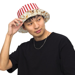 May include: A bucket hat with a popcorn design. The hat features a white and red striped top and a popcorn-printed brim. The hat is worn by a person wearing a black t-shirt.